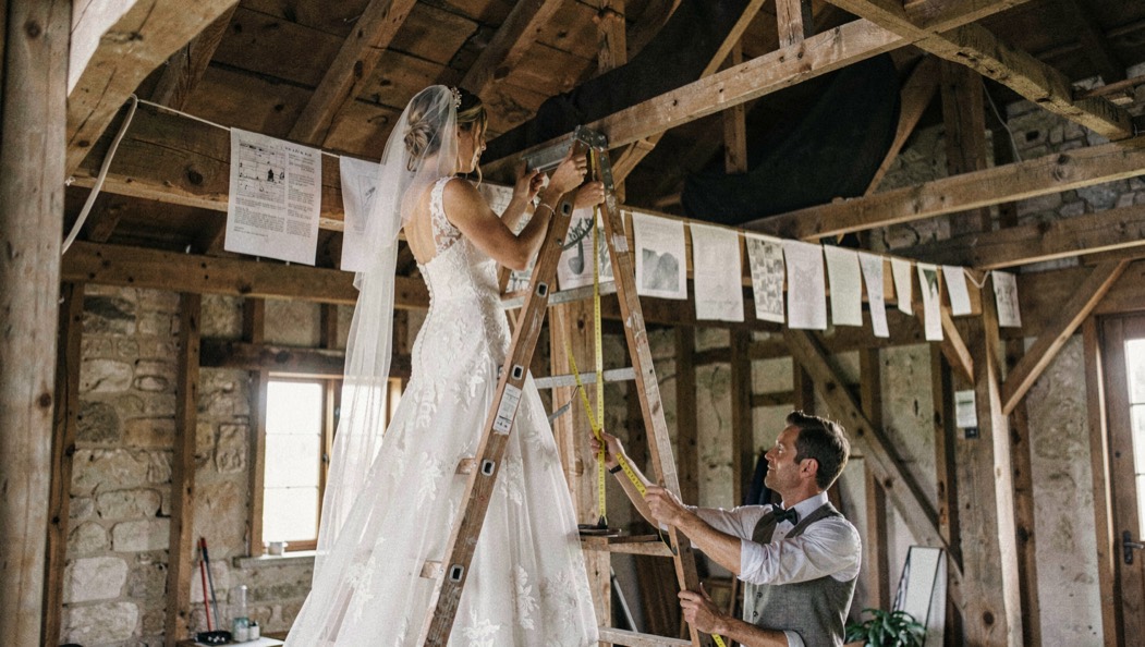 Bride on a wooden stepladder pinning paper banners between barn beams while a planner holds the tape measure below