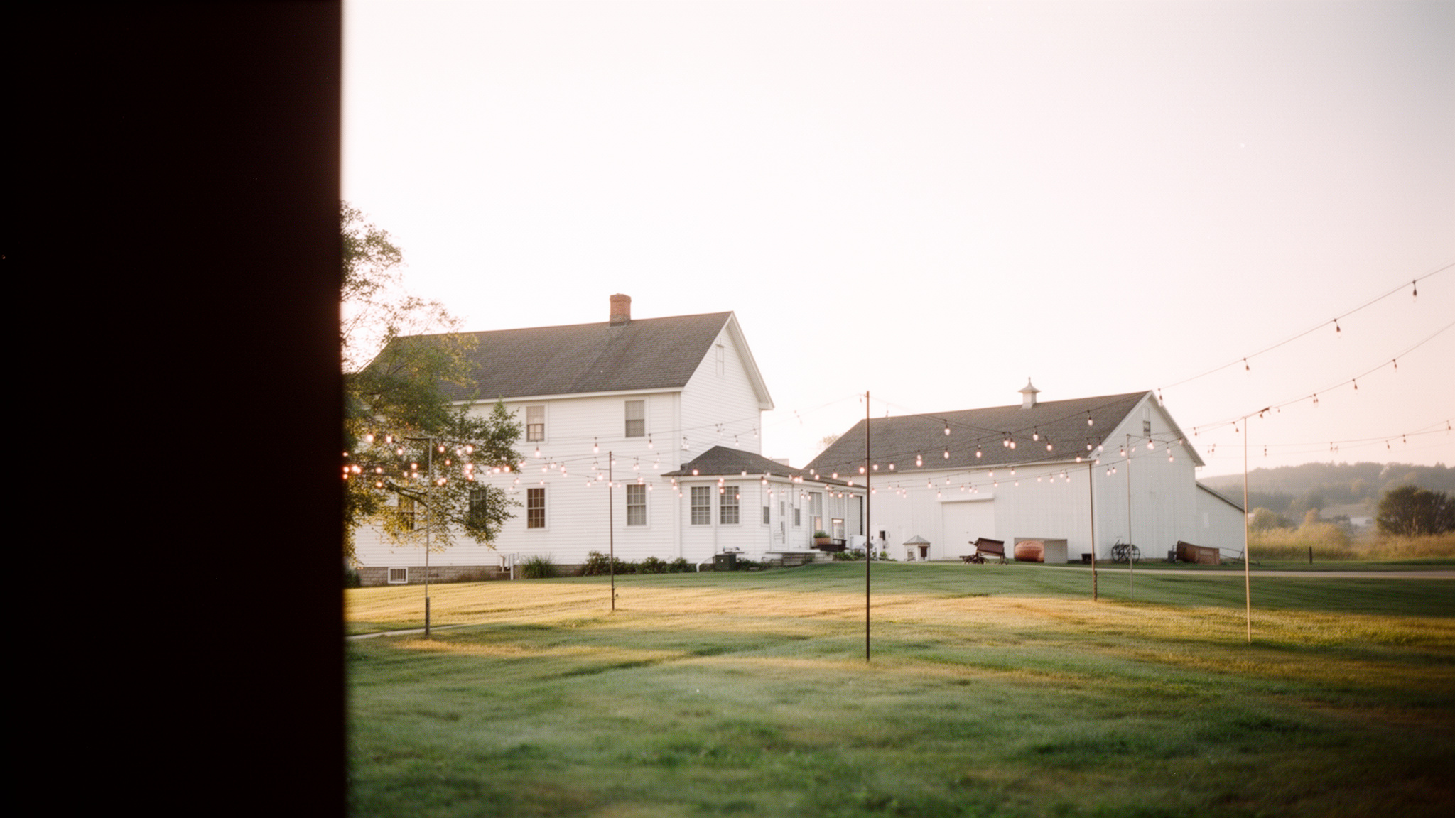 White clapboard farmhouse at golden hour with a wraparound porch, string lights strung from the posts and a mown lane winding toward the barn