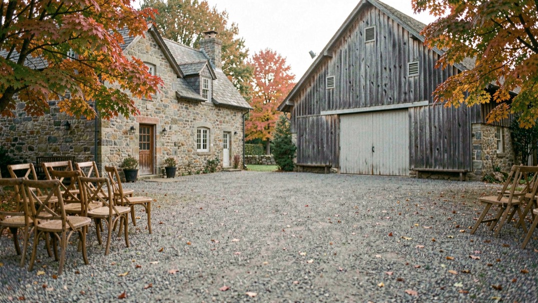 Gravel courtyard between a stone farmhouse and its converted dairy barn, wooden chairs stacked for setup and maple trees turning at the edges